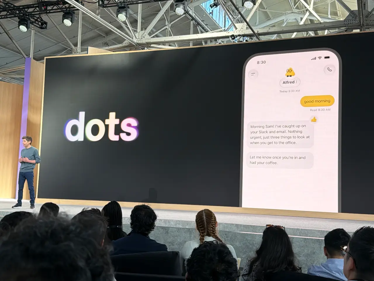演讲者站在舞台上，旁边的大屏幕显示“dots”标志和名为 Alfred 的助手的模拟手机对话。消息内容为：“早上好”、“早安 Sam！我已经查看了你的 Slack 和邮件，没有紧急事项，你到办公室后只需处理这三件事”，以及“到你到岗并喝完咖啡后告诉我一声。”